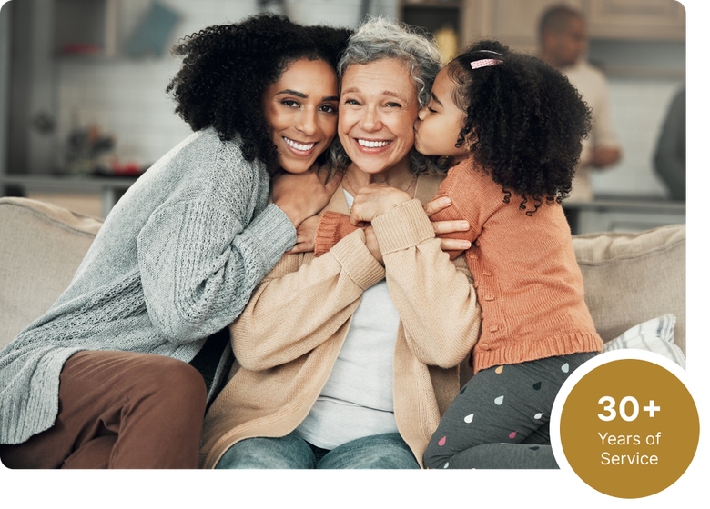 Three women hugging on a couch, smiling in a cozy living room, with a “30+ years” badge in the corner