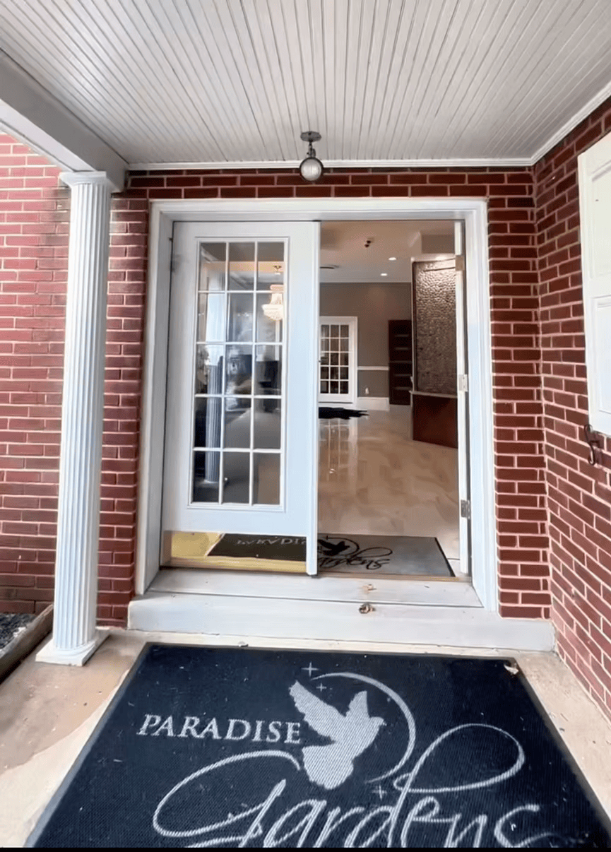 Front entry of a brick building with open glass doors and a black Paradise Garden welcome mat.