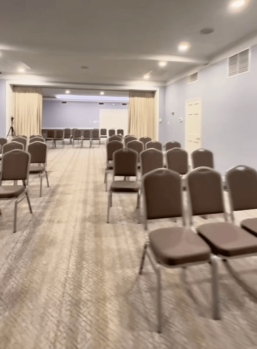 Empty conference room with rows of brown chairs facing a small stage and white projection screen.