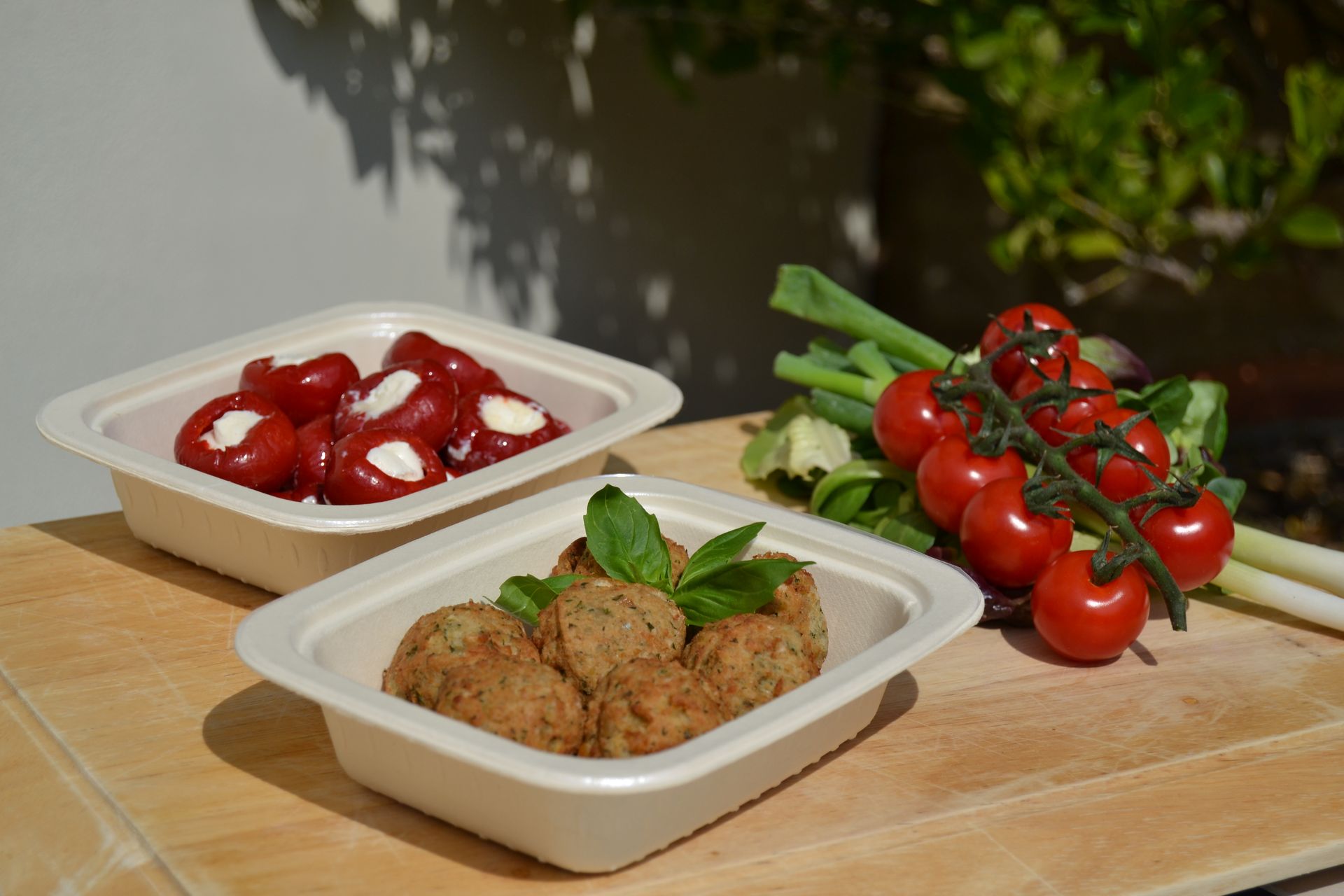 Two square bowls on a wooden board: one with red stuffed peppers, the other with meatballs and a sprig of fresh basil.