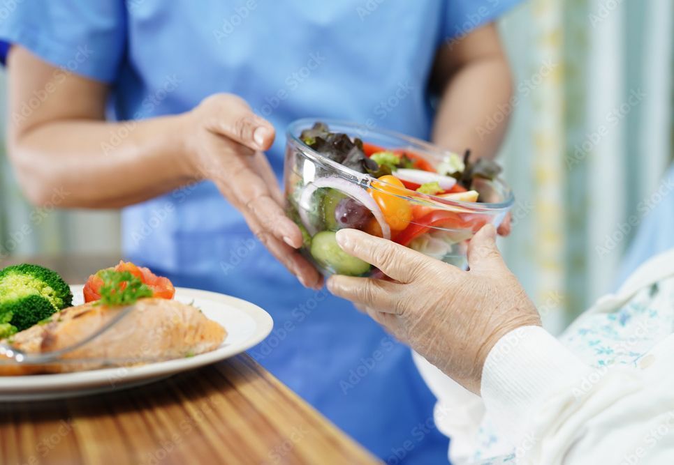 A healthcare worker in scrubs hands a bowl of fresh salad to a person, with a salmon dish on a table nearby.