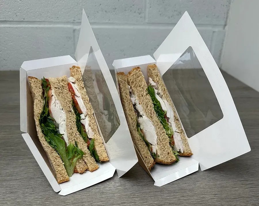 Two fresh chicken and lettuce sandwiches in individual white cardboard display boxes on a wooden surface.