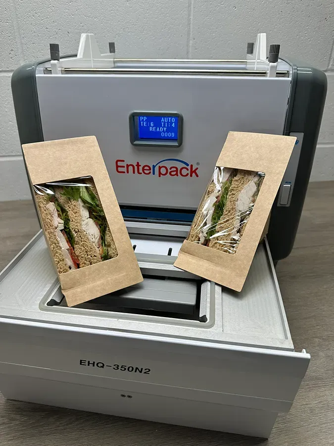 Two packaged sandwiches in kraft paper sleeves sit in front of an Enterpack food sealing machine.