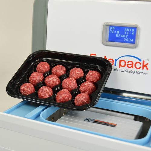 A black tray of raw meatballs held over an open Foodpack tray sealing machine with a blue digital display.