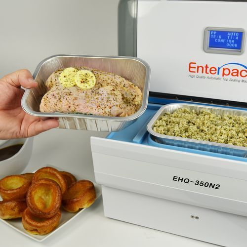 A person places a tray of seasoned chicken into an EHQ-350N2 Enterpack sealing machine next to cooked Yorkshire puddings.