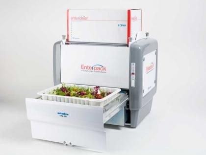 A white Enterpack food tray sealing machine with a box of film on top and a tray of salad inside the open drawer.