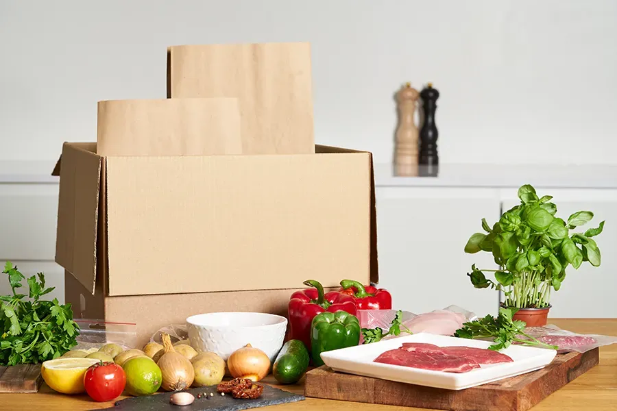 A cardboard delivery box with paper bags, raw meat, fresh vegetables, and herbs on a kitchen table.