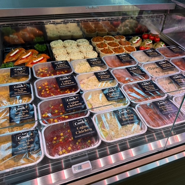 A refrigerated deli display case filled with rows of pre-packaged prepared meals, including chicken dishes and meatballs.