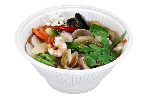A white bowl filled with spicy seafood soup containing shrimp, clams, mussels, enoki mushrooms, and fresh herbs.