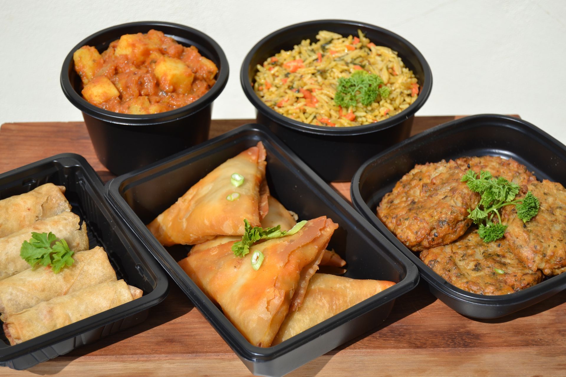 Takeout meal with curry, rice, spring rolls, samosas, and vegetable fritters in black containers on a wooden surface.