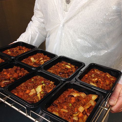 A person wearing a plastic apron holds a tray of black containers filled with a reddish-brown meat and vegetable stew.