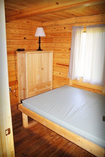 A bedroom in a log cabin with a bed , dresser , lamp and window.