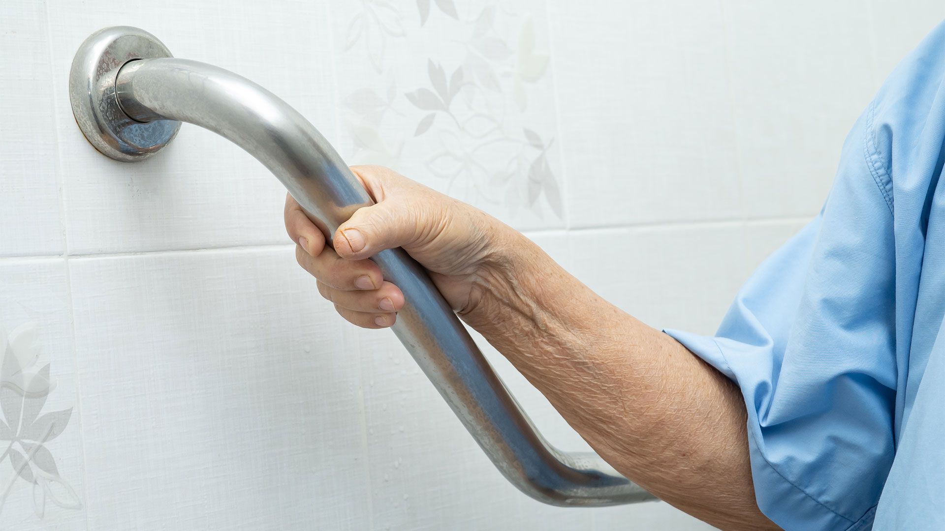 An elderly woman is holding onto a stainless steel hand rail in a bathroom.