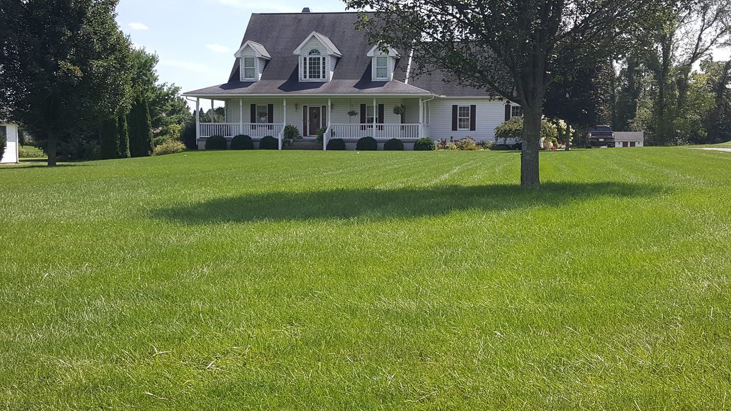A large white house sits in the middle of a lush green field White house with porch, dormers, and green lawn under a blue sky, trees surrounding.