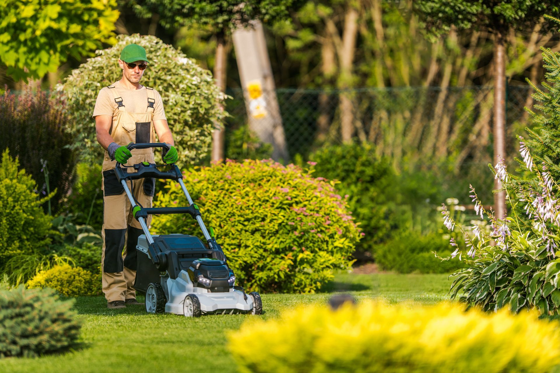 On a sunny day, a man in sunglasses mows the lawn, demonstrating effective lawn care solutions. Man mowing lawn in a sunny garden, wearing green hat, tan overalls, and gloves.