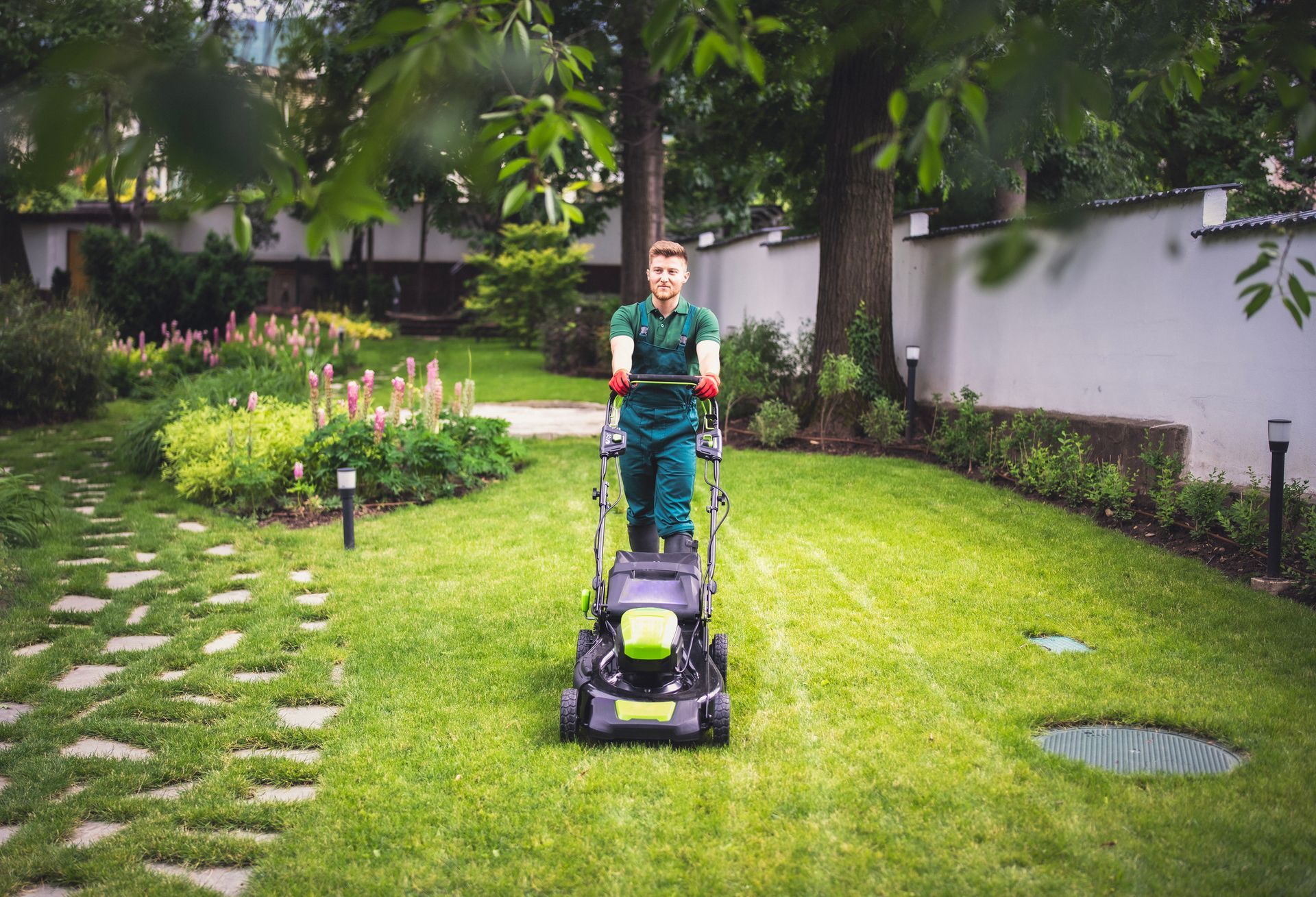 A man is mowing the lawn with a lawn mower on a sunny day, showcasing effective lawn care solutions. Man in green work clothes mowing a green lawn in a well-manicured garden.
