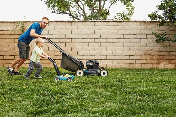 Father and child mowing the lawn together. The child has a toy mower.