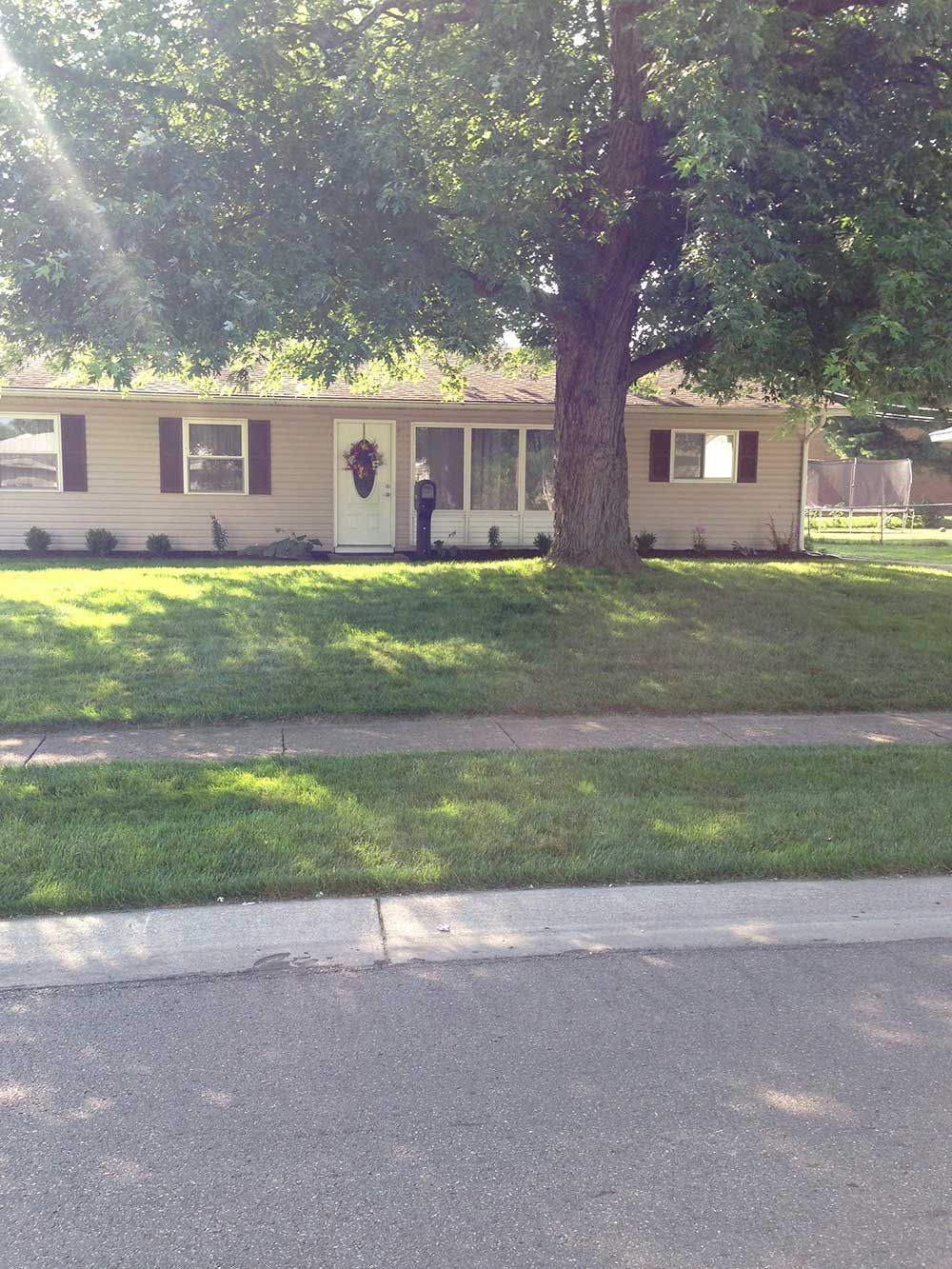Ranch-style house with beige siding, a large tree in front, and green grass. A person is standing near the front door.