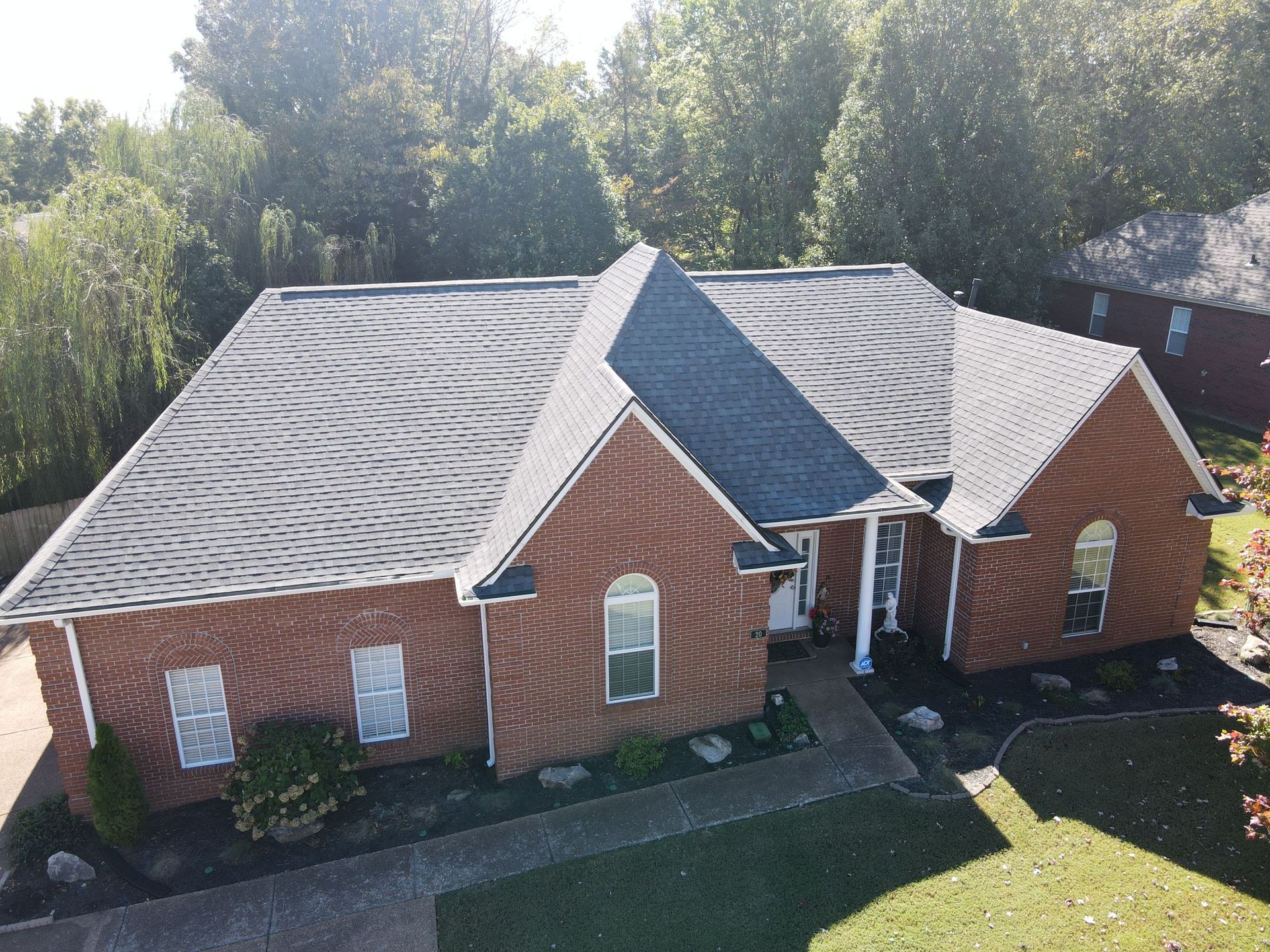 Brick house with gray shingle roof and dormer, seen from above amid trees and lawn