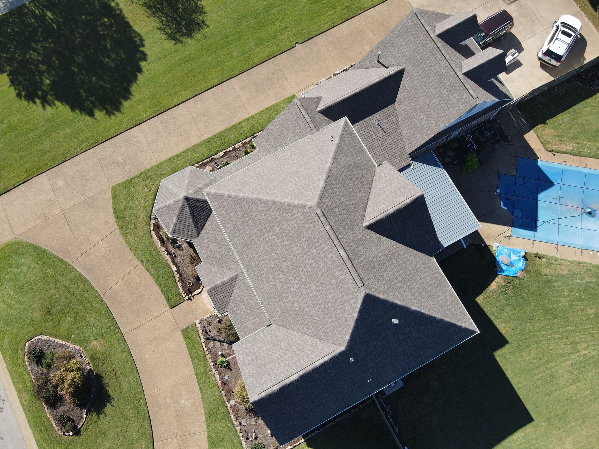 Aerial view of a gray-roofed house beside a driveway, lawn, and blue-covered pool.