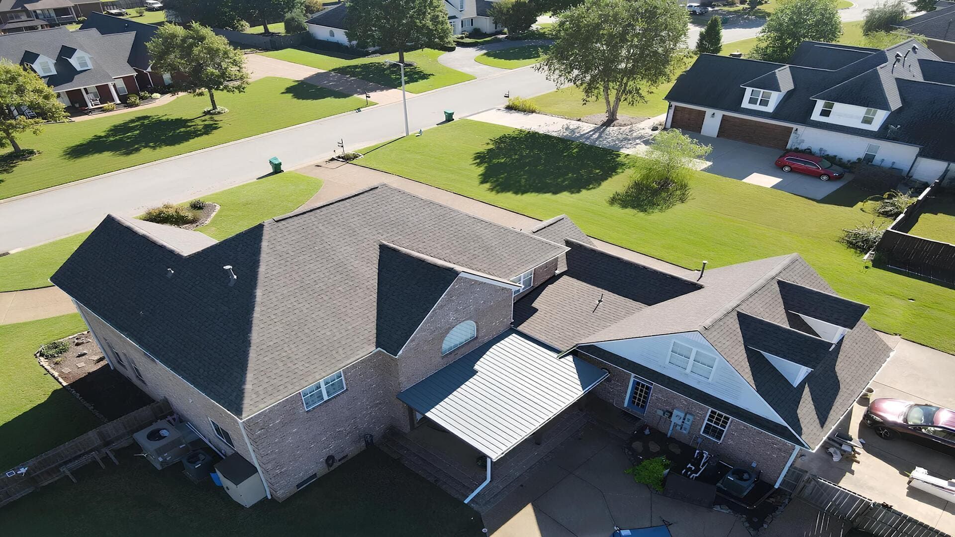 Aerial view of a suburban house with dark roof, driveway, and neighboring homes on a sunny day