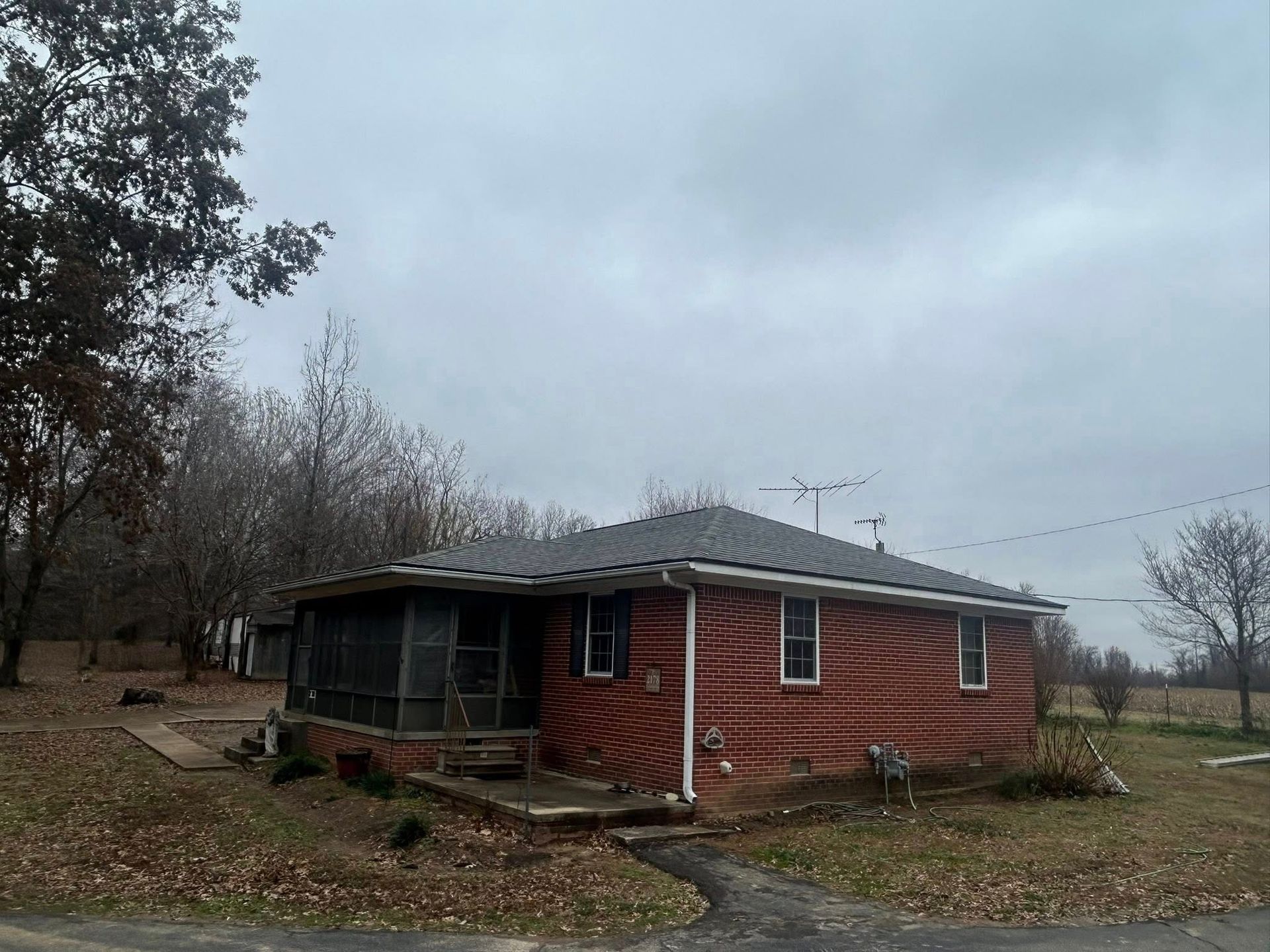 Small red brick house with a screened porch on a cloudy day, surrounded by bare trees and grass.
