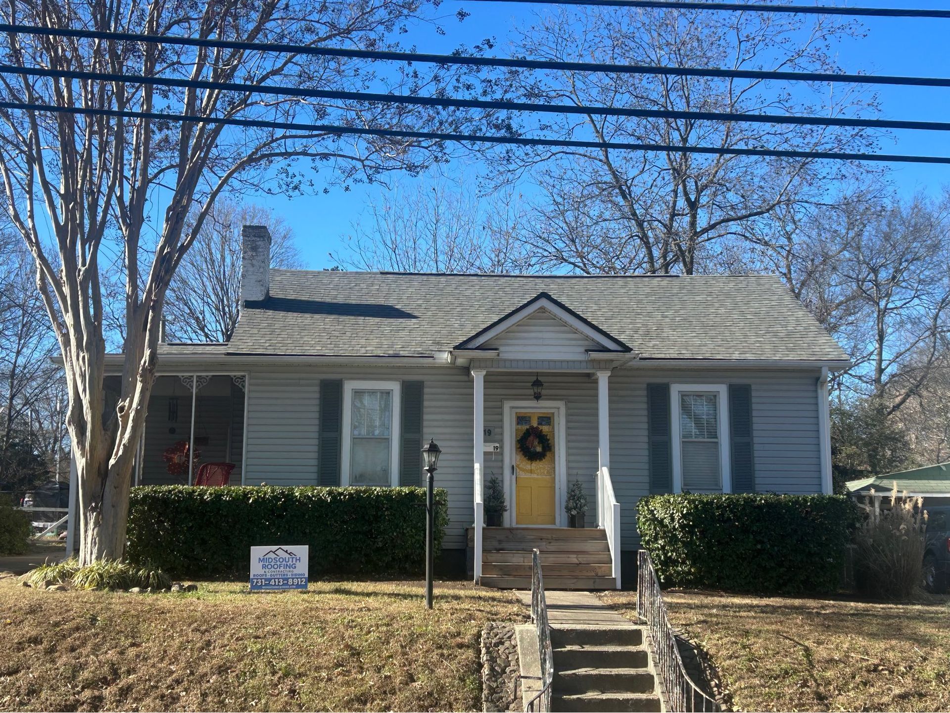 Small gray house with white trim, front porch, steps, and a hedge-lined yard under a blue sky.