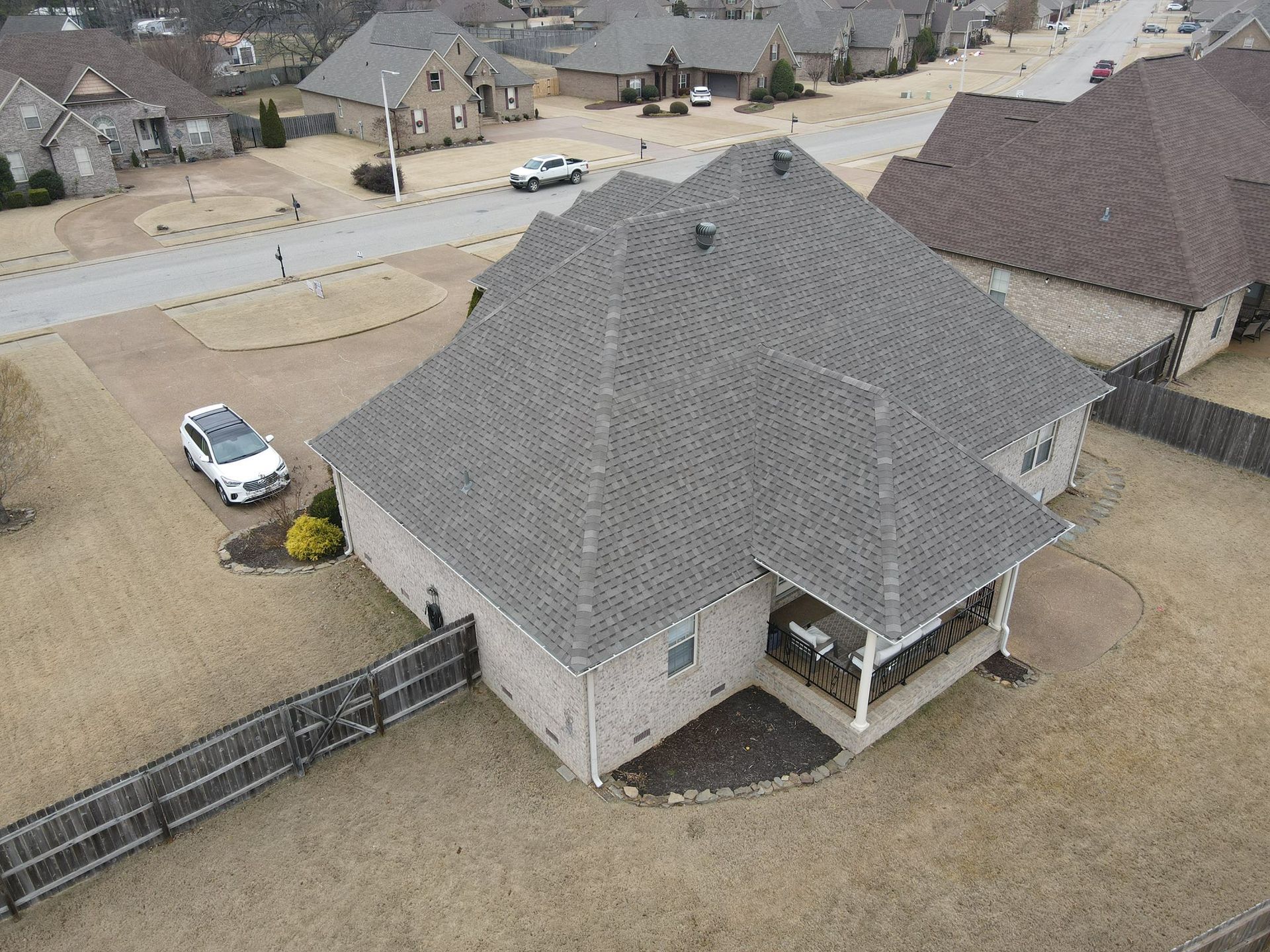 Aerial view of a gray house with a covered porch, fenced yard, and parked white SUV in a suburban neighborhood.