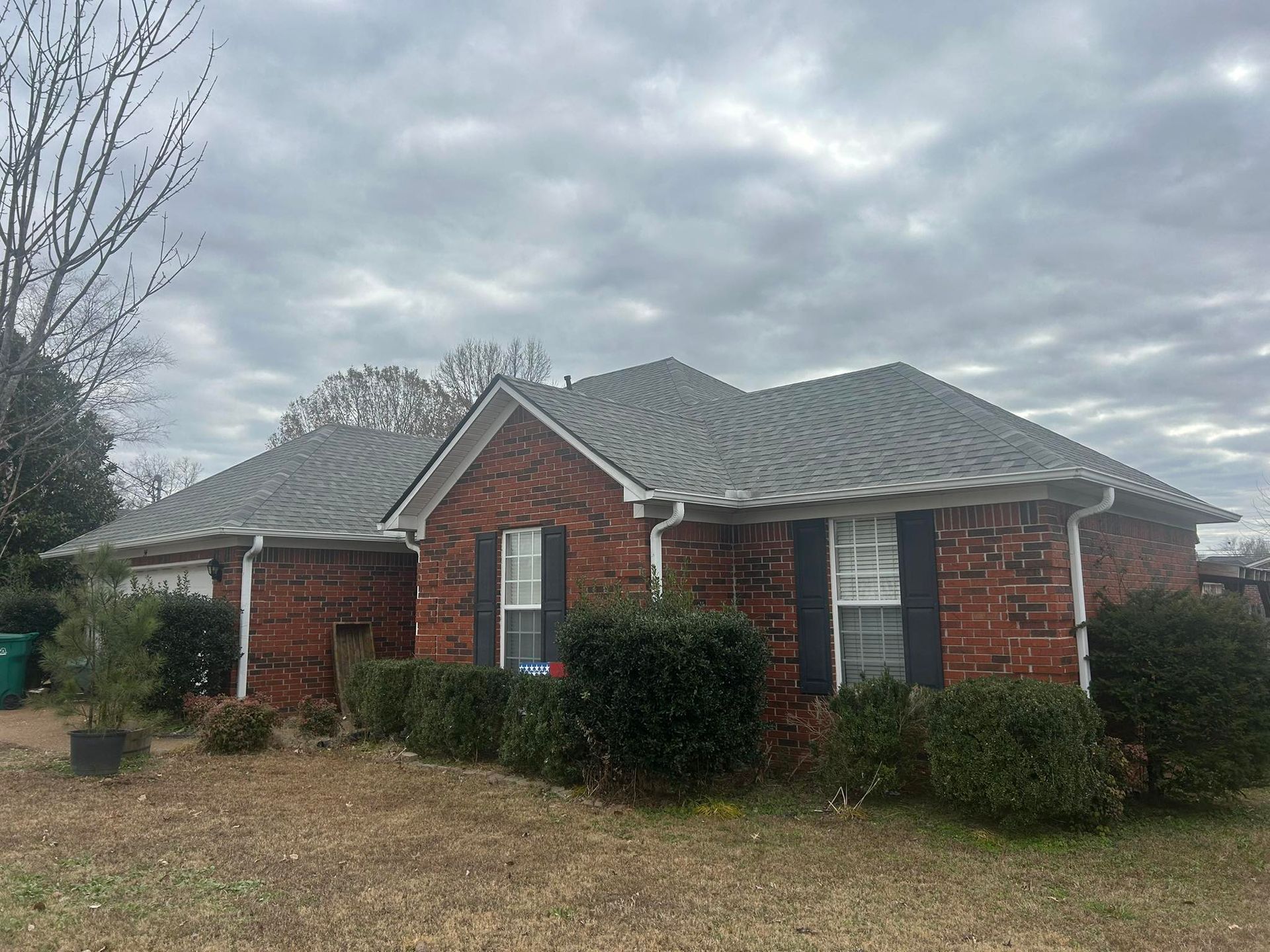 Single-story red brick house with gray shingled roof and shrubs under a cloudy sky