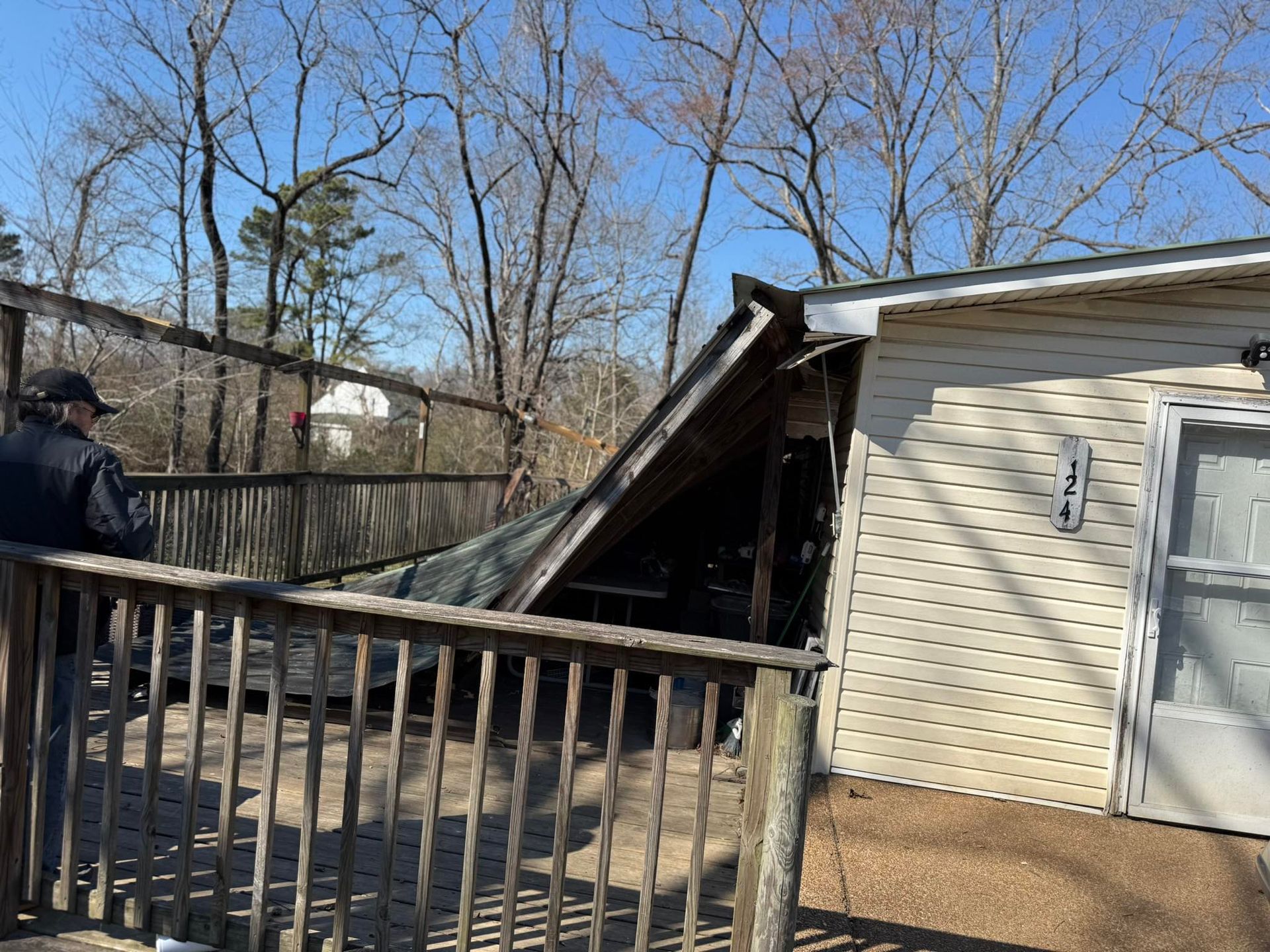 Collapsed deck attached to a house beside a wooden fence, with debris leaning into the yard.