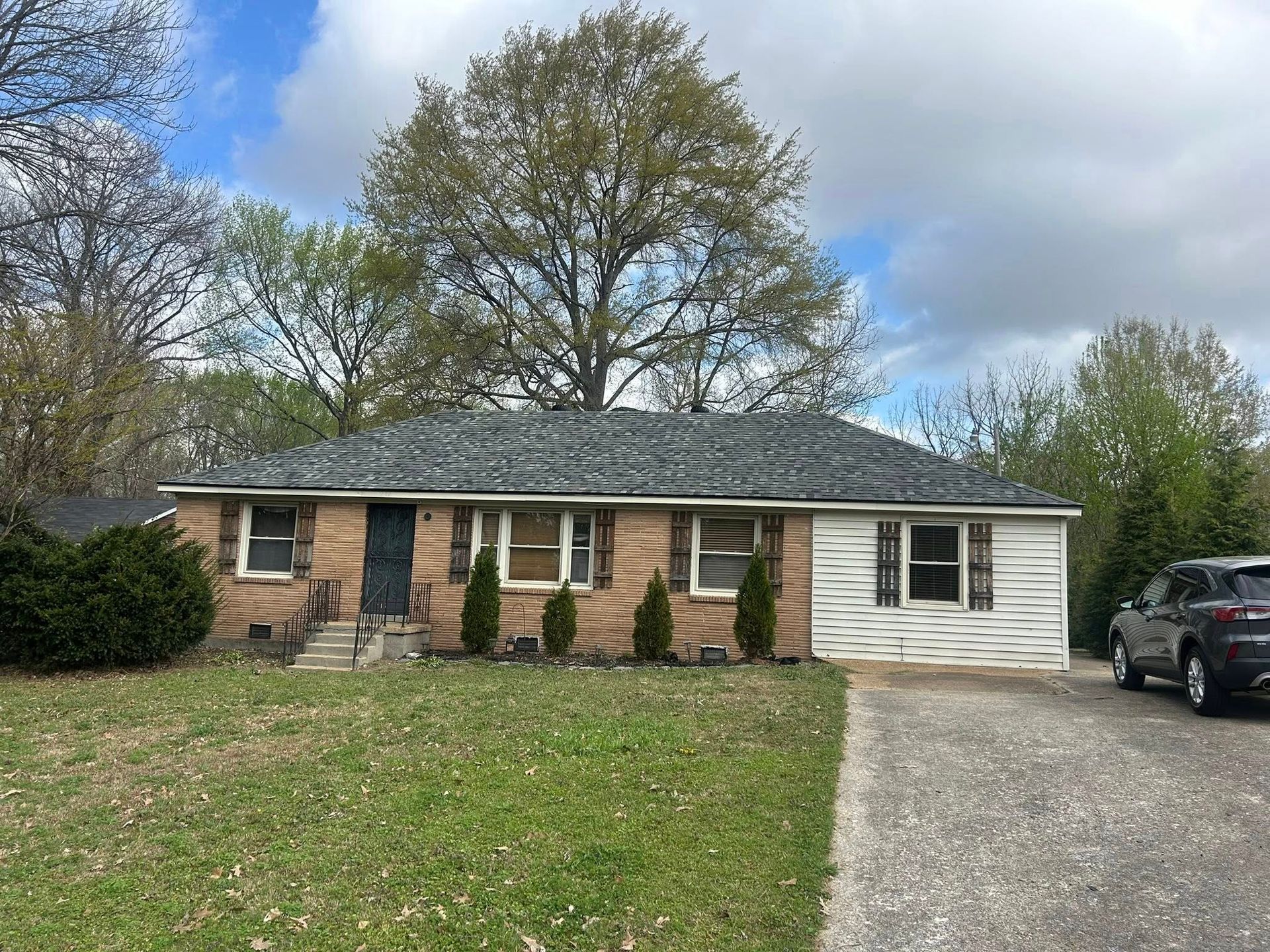 Single-story brick house with white siding, gravel driveway, and parked car under cloudy sky
