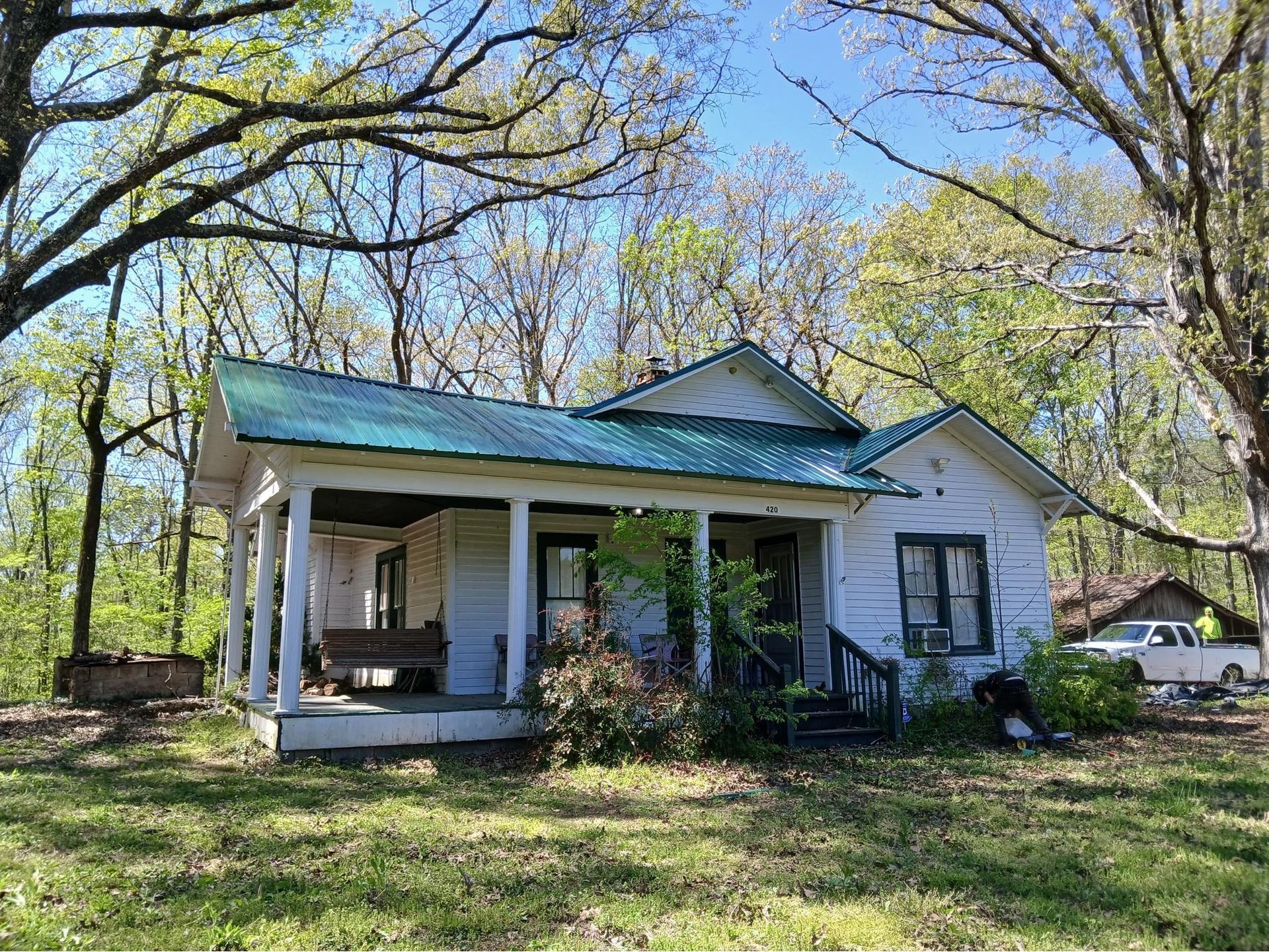 Small white house with a teal roof and front porch, surrounded by trees and grass.