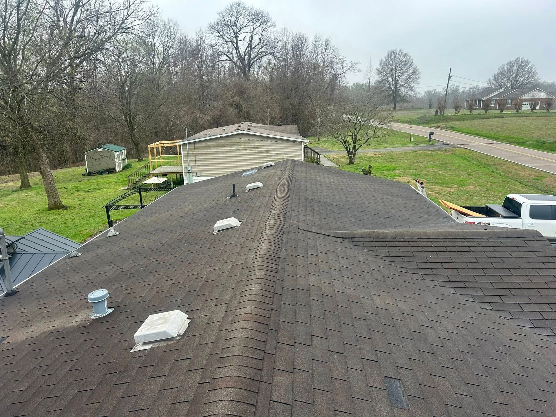 Roof of a house with dark shingles, vents, and a rural yard with trees and a road nearby.