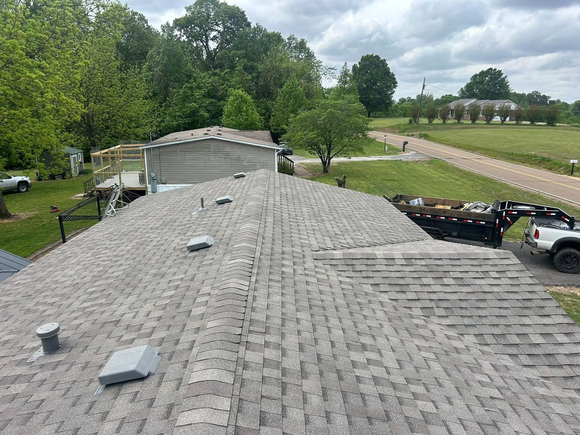 Rooftop view over gray shingles with trees, gravel drive, and parked vehicles beside a rural building