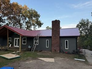 Dark green house with a red roof and brick chimney, with a ladder leaning against it and trees around it