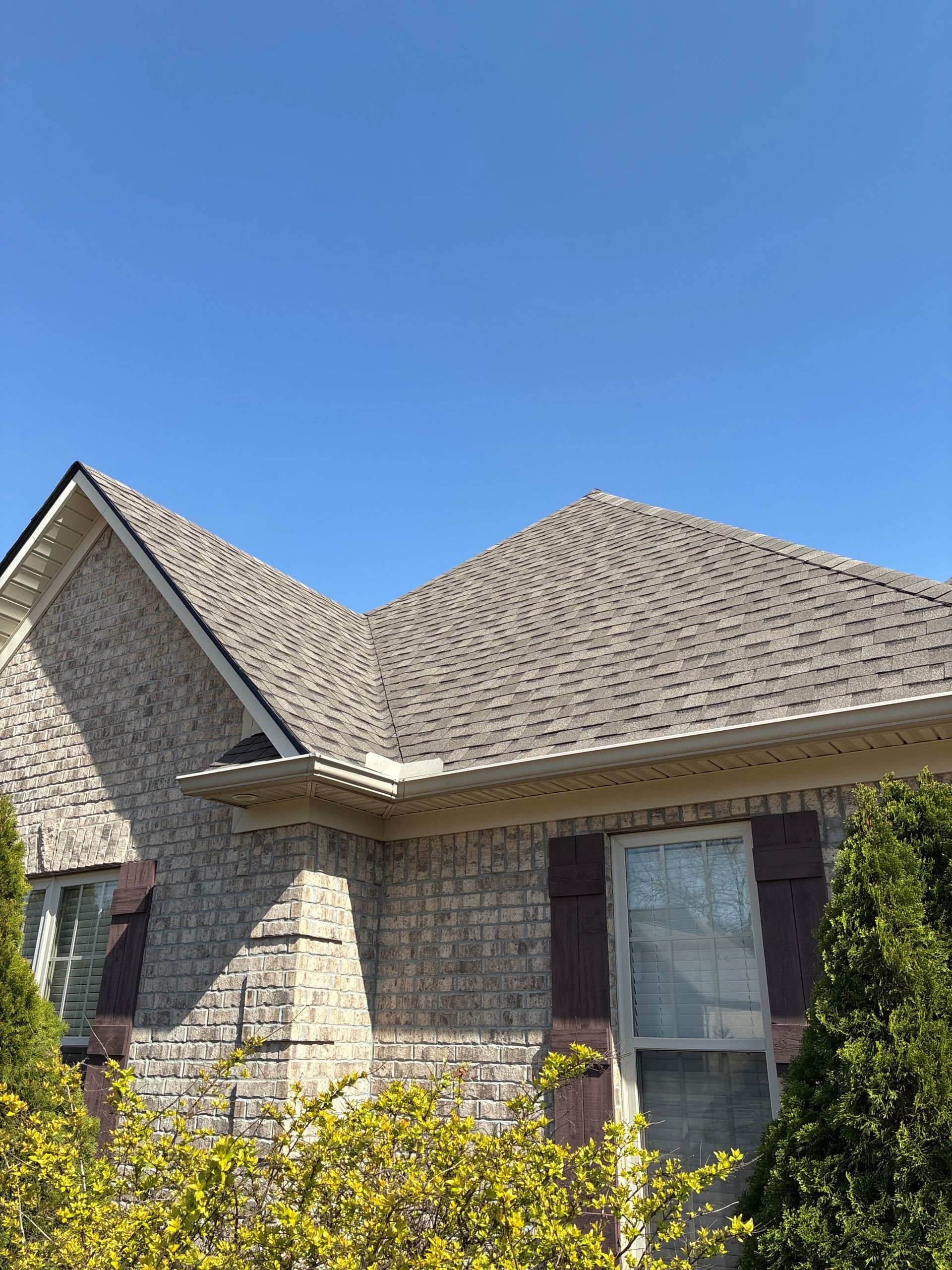 Brick house exterior with tan shingle roof, white windows, and shrubs under a clear blue sky