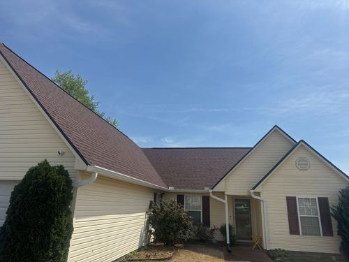 Suburban house exterior with beige siding, brown roof, and a small front porch under a blue sky