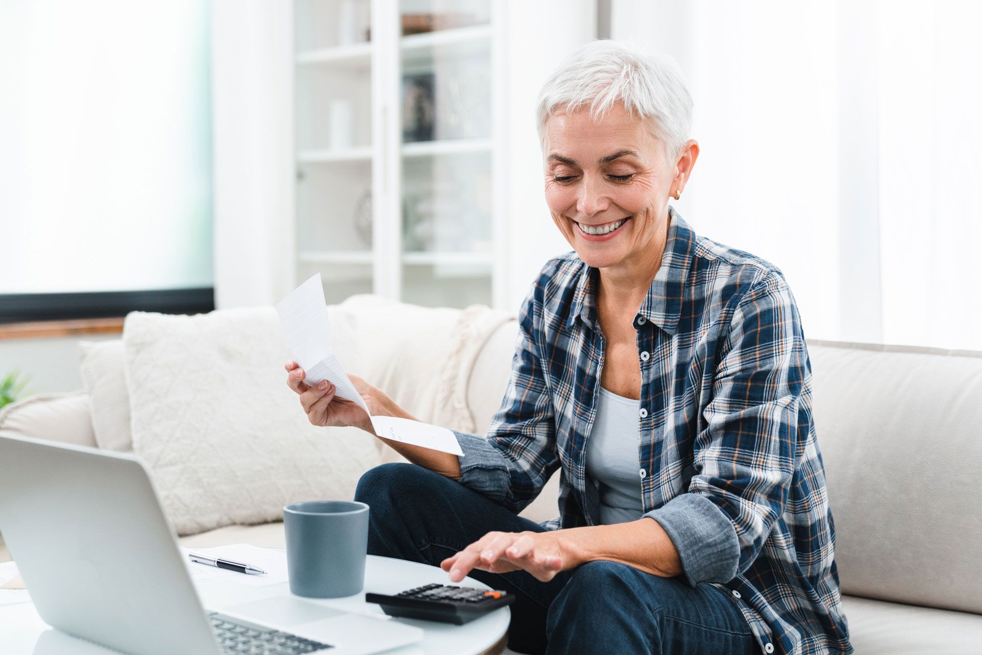 Woman sits on couch, reviewing bills with calculator and laptop. Smiling, in plaid shirt.