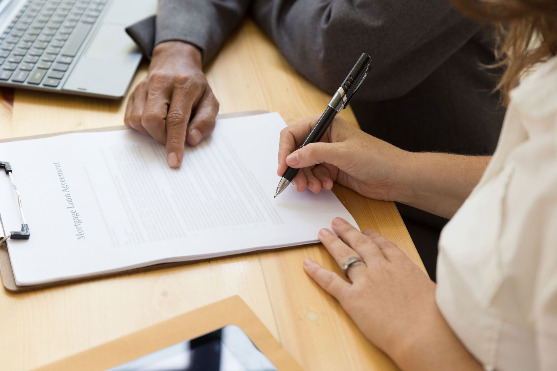 A person signing a document on a clipboard while another points to a spot; laptop visible.