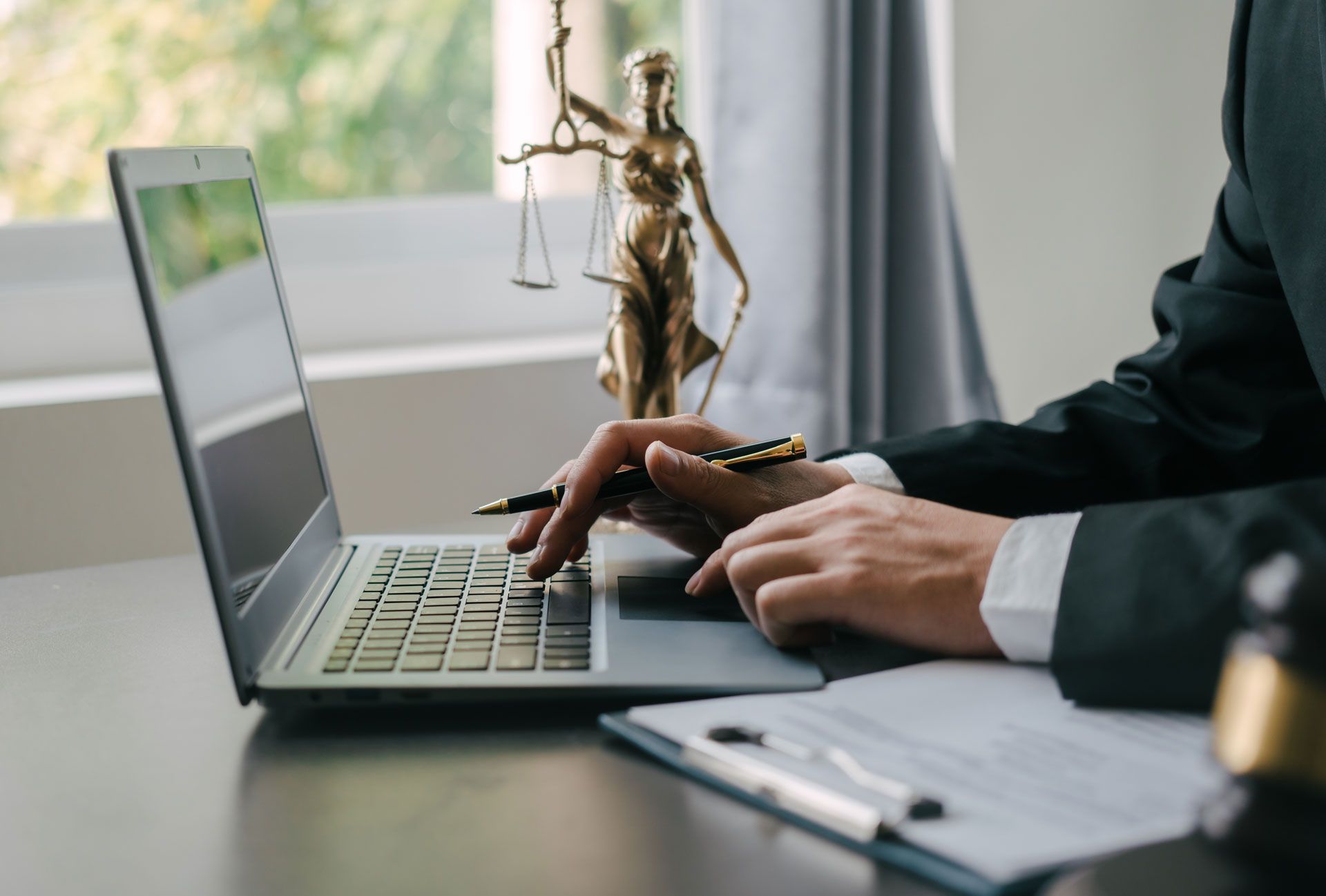 Person in suit typing on a laptop with a Lady Justice statue and documents nearby.
