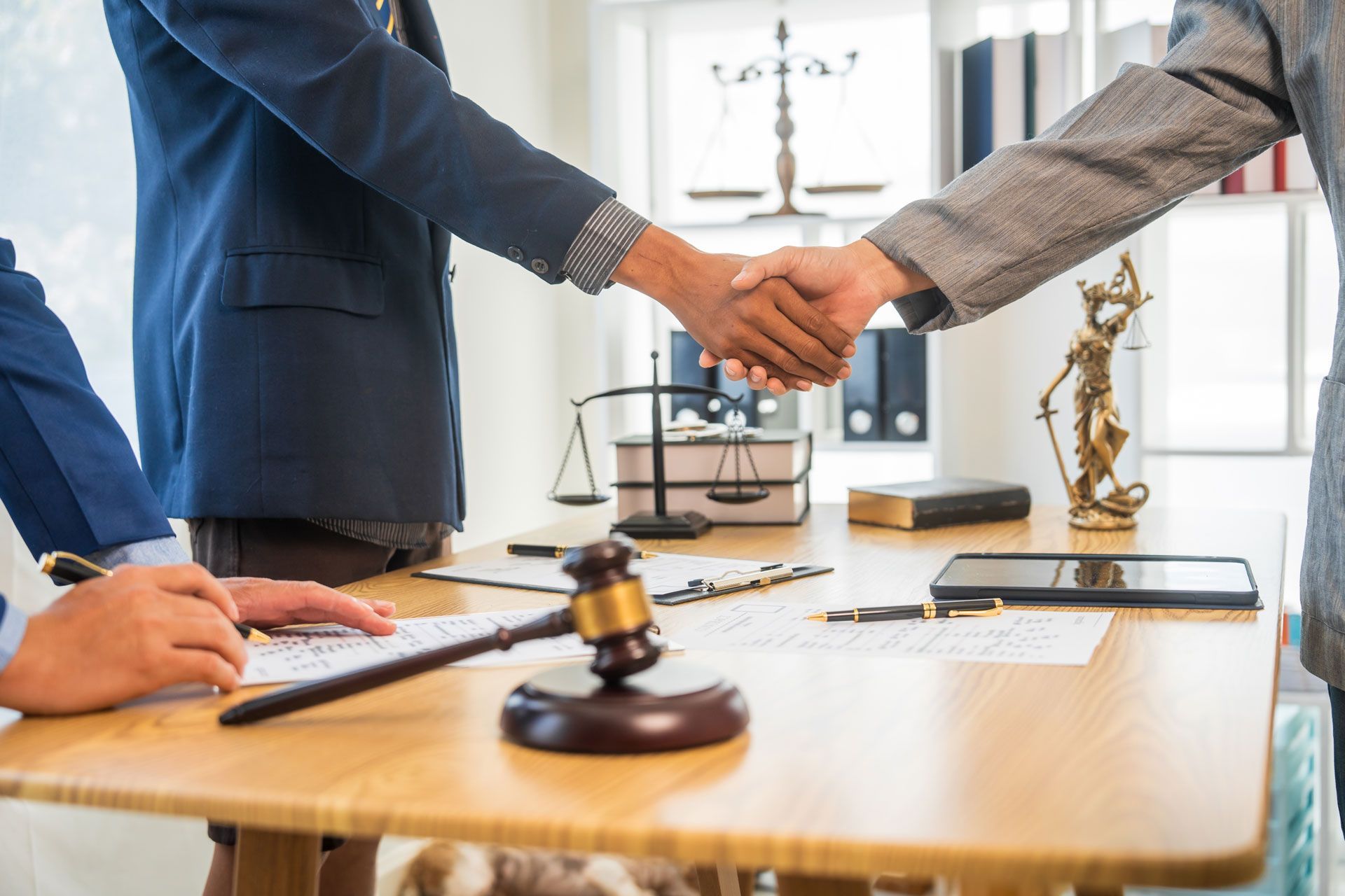 Two people shaking hands over a table with legal documents, scales, and a gavel.