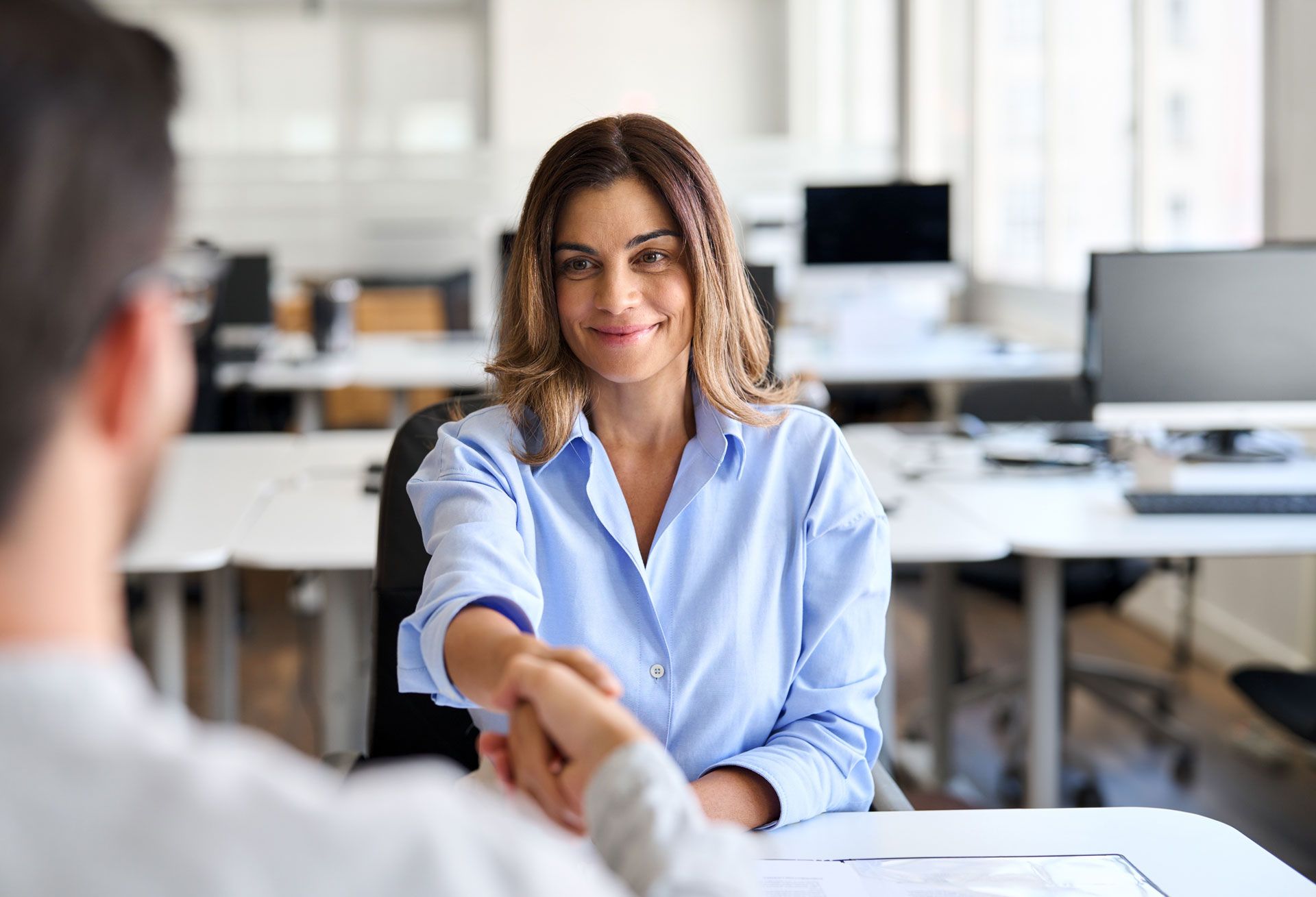 Woman in blue shirt shaking hands with a person, smiling in an office setting.