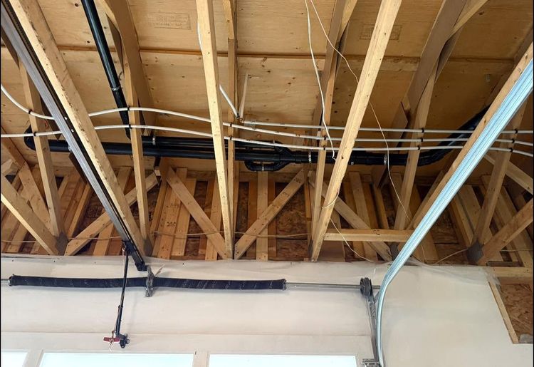 Open ceiling with exposed wooden trusses, pipes, and ductwork above a white wall.