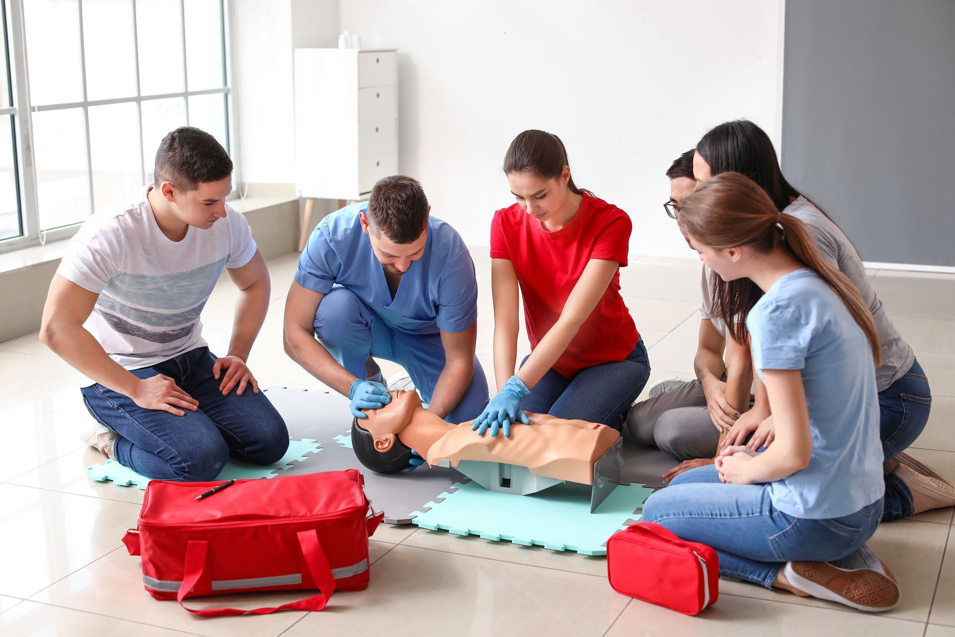 People in a first aid training course practicing CPR on a mannequin, indoors. A red medical bag sits in front.