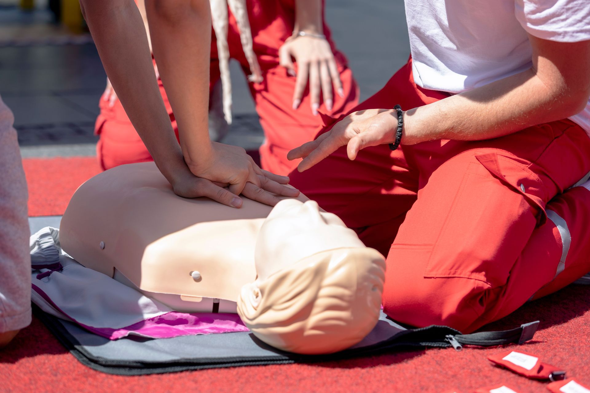 People practicing CPR on a training dummy, wearing red pants and white shirts outdoors.