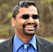 Man with sunglasses and beard smiling, wearing a suit jacket and blue shirt.