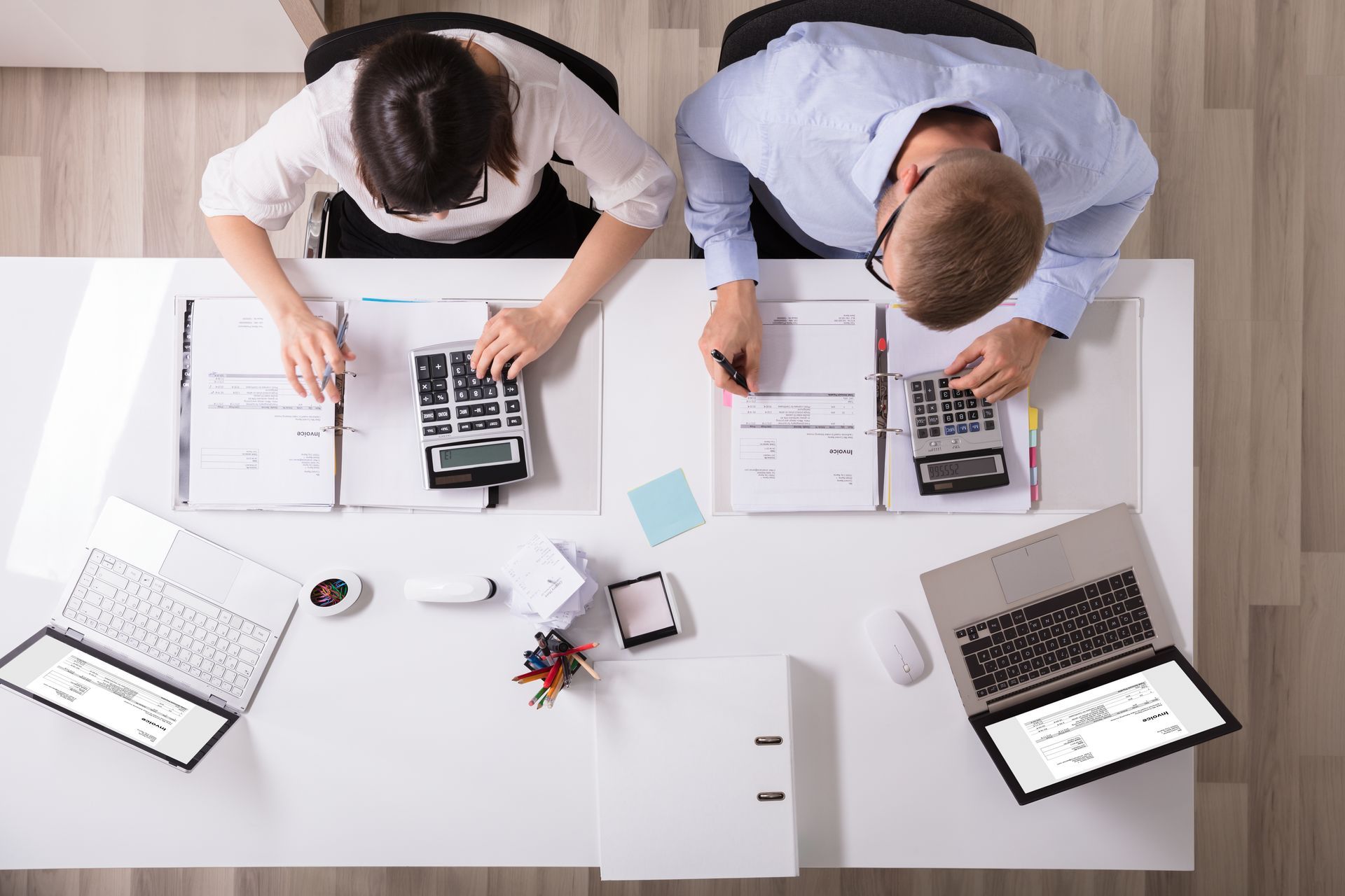 Two people at a white desk, calculating using calculators and laptops, reviewing papers.