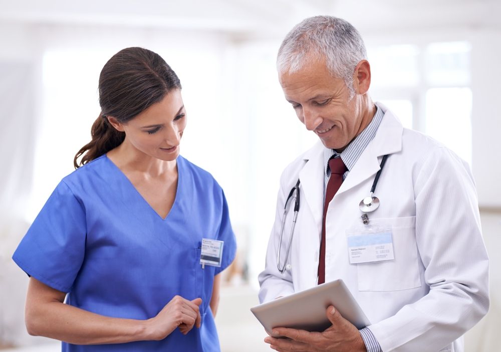 Nurse and doctor looking at a tablet, indoors. The nurse wears blue scrubs; the doctor, a white coat and stethoscope.