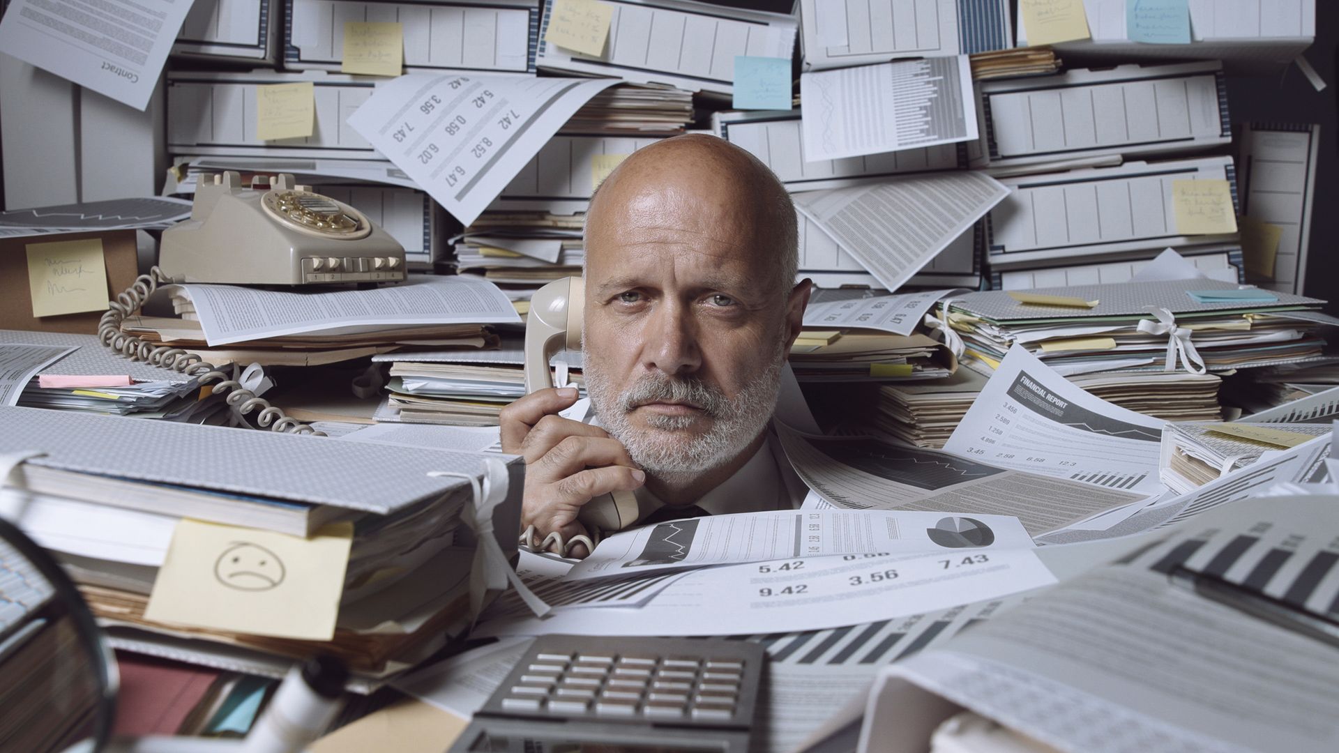 Bald man surrounded by paperwork, on the phone, looking stressed.