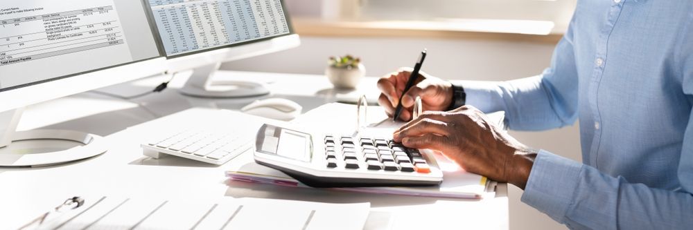 Person calculates with a calculator and pen at a desk, with a computer and papers.